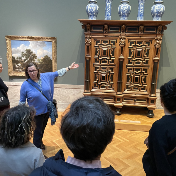 Dr. Claudia Swan stands facing the group with a large cabinet topped with five porcelain vases behind her.