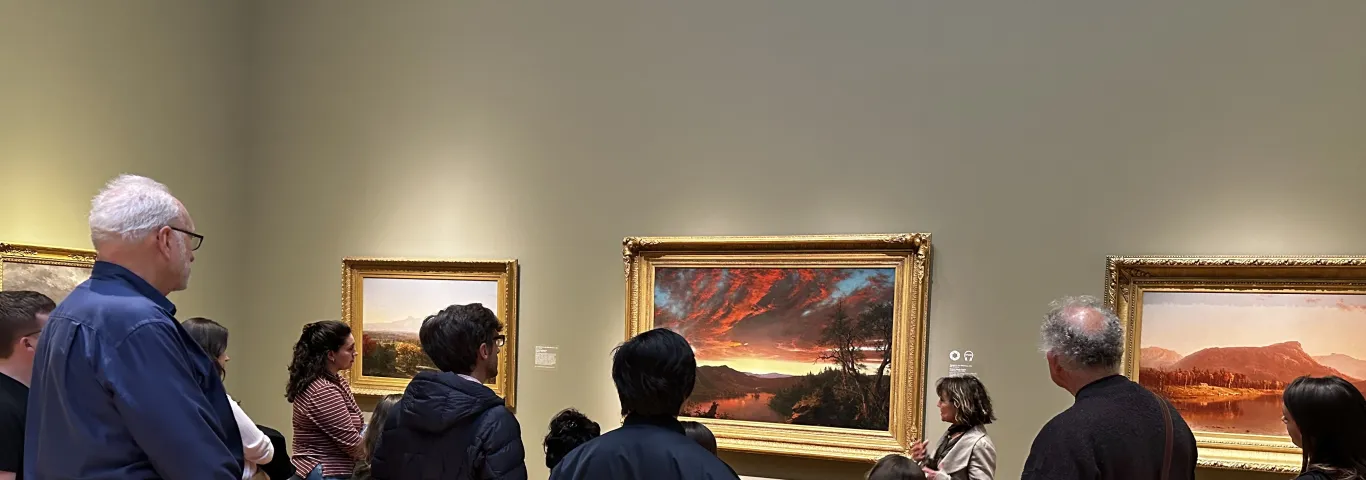 An image showing a group of students and faculty looking at Dr. Angela Miller, who is standing in front of Frederic Edwin Church's painting Twilight in the Wilderness